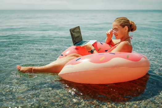 Woman laptop sea. Freelancer woman in sunglases floating on an inflatable big pink donut with a laptop in the sea. People summer vacation rest lifestyle concept