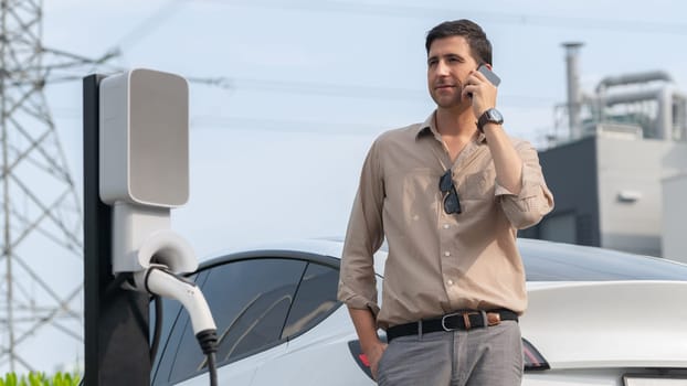 Man talking on the phone while recharge EV car battery at charging station connected to power grid tower electrical as electrical industry for eco friendly car utilization. Panorama Expedient