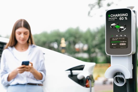 Young woman using smartphone online banking application to pay for electric car battery charging from EV charging station during vacation holiday road trip at national park or summer forest. Exalt