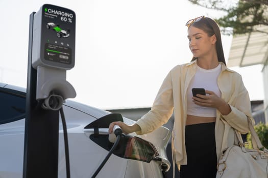 Young woman holding shopping bag and use smartphone to pay for electricity for recharging EV car battery from charging station at city mall parking lot. Modern woman go shopping by eco car. Expedient