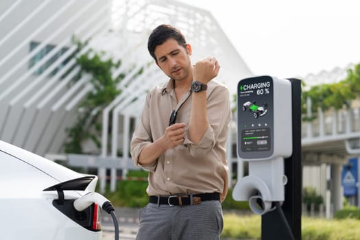 Young man checking time on smartwatch while EV charger to recharging battery from charging station in city mall parking lot. Rechargeable EV car for sustainable eco friendly urban travel. Expedient