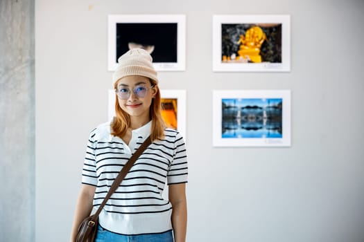 Visitor woman smiling on art gallery collection in front framed paintings pictures on white wall, lifestyle Asian people watch at photo frame to leaning against at show exhibition artwork gallery