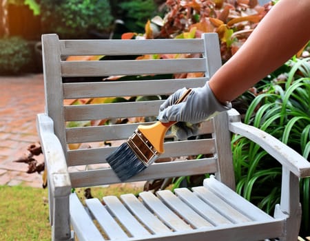 Garden chair with brush in protective gloves. Worker paints garden furniture. Renewal, renovation of wooden garden furniture