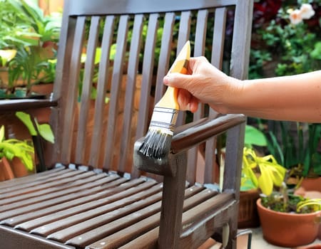 Garden chair with brush in protective gloves. Worker paints garden furniture. Renewal, renovation of wooden garden furniture