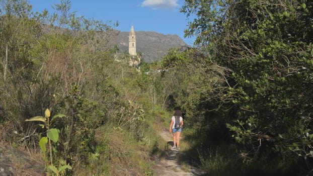 Hiker treks on forest path, church spire visible far.