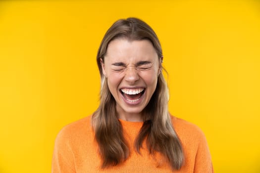 Happy emotions concept. Positive and beautiful young woman laughs poisitively looks aside with carefree face expression wears casual orange sweater isolated over yellow studio background.