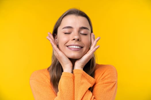 Happy emotions concept. Positive and beautiful young woman laughs poisitively looks aside with carefree face expression wears casual orange sweater isolated over yellow studio background.