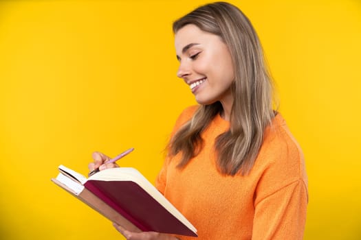 Happy emotions concept. Positive and beautiful young woman laughs poisitively looks aside with carefree face expression wears casual orange sweater isolated over yellow studio background.