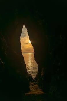 A cave with a small opening in it and the sun shining through it. The sun is setting and the water is calm