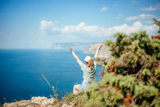A woman is sitting on a hill overlooking the ocean, with her arms raised in the air. Concept of freedom and joy, as the woman seems to be celebrating the beauty of nature and the vastness of the ocean