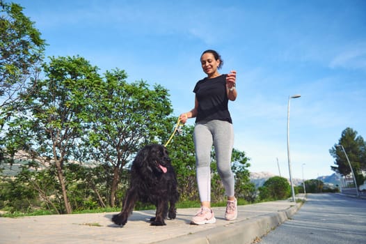 Full size happy multi ethnic woman walking her dog on leash in the nature. Smiling female in sportswear, enjoying walk with her pedigree cocker spaniel outdoors. People and domestic animals concept