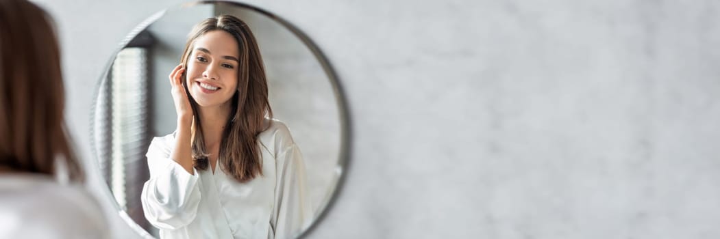 Beauty Concept. Portrait Of Attractive Happy Woman Looking At Mirror In Bathroom, Beautiful Millennial Lady Wearing White Silk Robe Smiling To Reflection, Enjoying Her Appearance, Copy Space