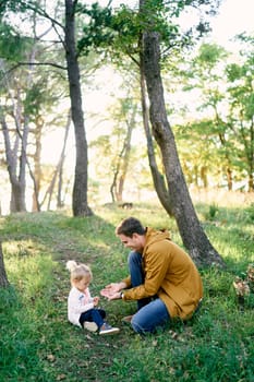Dad holds out mushrooms to a little girl sitting on green grass in the forest. High quality photo