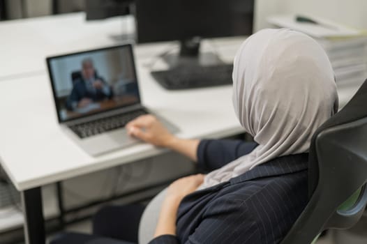 A pregnant woman in a hijab communicates with a colleague via video conference on a laptop