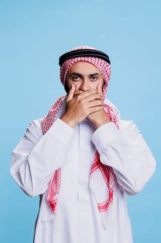 Arab man wearing traditional islamic clothes holding palms on mouth and asking to keep quiet portrait. Muslim person showing speak no evil three wise monkeys concept and looking at camera