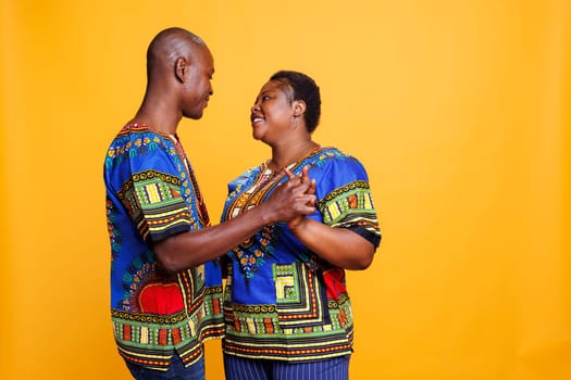 Happy african american spouse wearing ethnic clothes dancing slow dance and showing love. Mid adult man and woman romantic couple holding hands and looking in eyes with affection feeling
