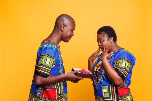 Excited african american woman receiving festive gift box from smiling man. Black boyfriend wearing ethnic clothes giving relationship anniversary present box to surprised girlfriend