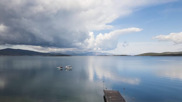 A natural landscape featuring a lake with a dock in the foreground, surrounded by water and a cloudy sky with cumulus clouds in the background, creating a picturesque horizon