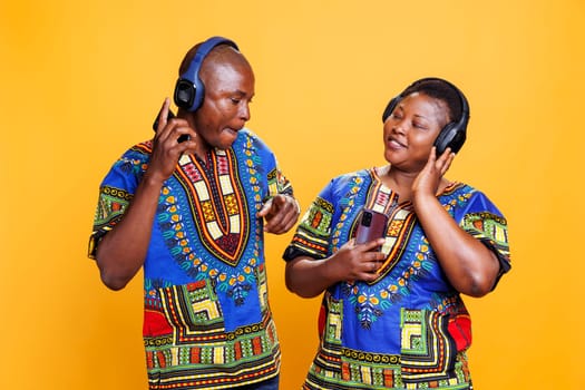Smiling black couple dressed in ethnic clothes listening to music in headphones. Joyful man in wireless earphones dancing while woman with cheerful expression holding smartphone