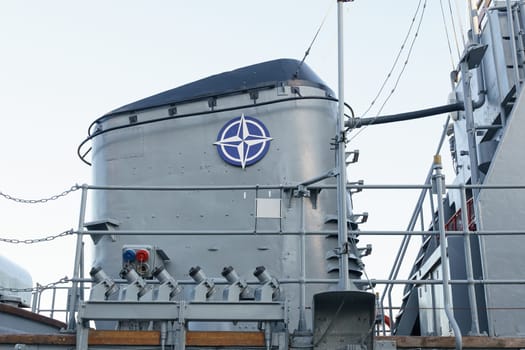 Klaipeda, Lithuania - August 11, 2023: A close-up view of a naval ships deck, showcasing a large gray gun turret with a NATO insignia, festooned with colorful signal flags under a clear sky.