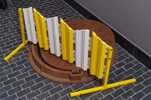A bright yellow foldable safety fence surrounds an open brown well hatch on a gray paved sidewalk, alerting pedestrians to the potential hazard.