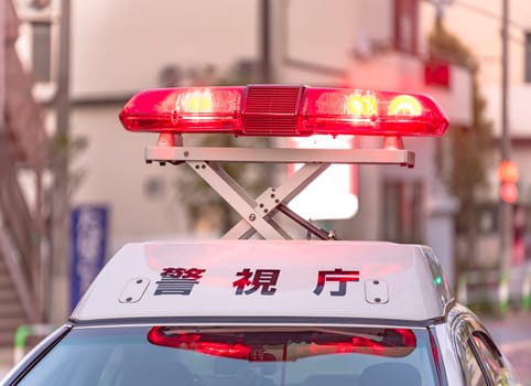 Close-up on bright red emergency beacons casting an intense glow as they swirl unfolded above the white roof of a Japanese police vehicle showing the word 'Keishicho' meaning 'Police'.