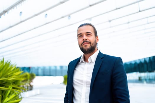 Successful businessman in suit with beard standing in front of office building confidently looking away. Hispanic male business person side view portrait, Free space. Modern business people concept