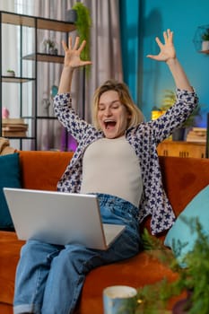 Surprised young woman using laptop, receive good news shocked by sudden victory celebrate lottery jackpot win in apartment. Cheerful girl with netbook sitting on sofa in living room at home. Vertical