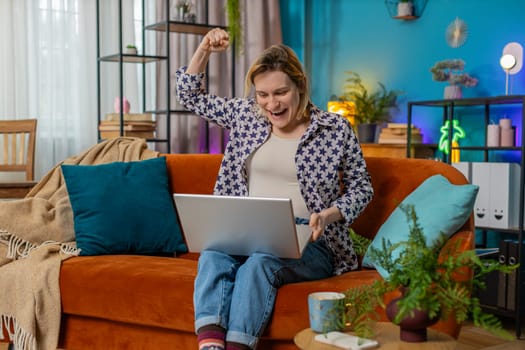Surprised Caucasian young woman using laptop, receive good news shocked by sudden victory celebrate lottery jackpot win in apartment. Cheerful girl with netbook sitting on sofa in living room at home.