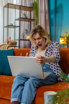 Displeased woman using laptop notebook typing browsing working, loses becoming surprised sudden lottery results bad news fortune loss game fail computer virus. Young girl at home on couch. Vertical