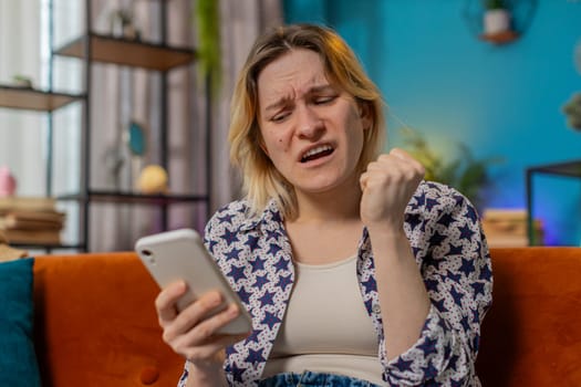 Shocked sad frustrated woman using smartphone reading negative message feels annoyed sitting on sofa in living room at home. Upset Caucasian girl having gadget trouble in apartment. App crash concept