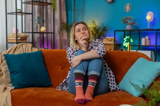 Sad lonely Caucasian woman looks pensive thinks over life concerns suffers from unfair situation sitting on sofa. Upset girl lean on hand depressed feeling bad annoyed in living room at home.