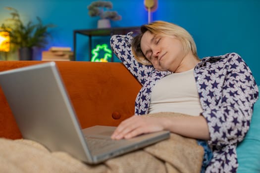 Bored sleepy young Caucasian woman working on laptop computer, leaning on hand sitting on couch with blanket. Exhausted tired freelancer workaholic girl sleeping in living room at home apartments