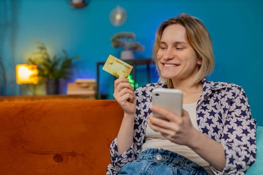Happy excited woman doing payments online purchase shopping in internet store. Smiling Caucasian girl holding banking credit card involved in verification process in mobile application on sofa at home