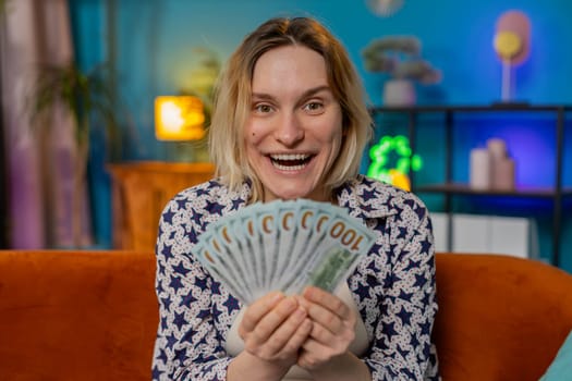 Successful rich blonde woman holding waving dollar bill money fan sitting on couch at home room. Happy excited girl winning online casino lottery game planning vacation calculating budget at home.