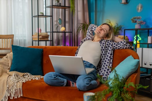 Happy young relaxed woman sitting on couch after finishing laptop work in living room. Caucasian girl student working online, remote job at home sofa. E-learning, browsing internet on netbook.