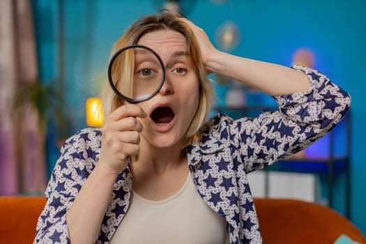 Investigator researcher scientist Caucasian woman holding magnifying glass near face looking into camera with big zoomed funny eye searching analyzing. Surprised amazed girl sitting at home room couch