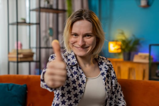 Portrait of excited smiling Caucasian woman looking approvingly at camera showing double thumbs up, like sign, good news, positive feedback. Happy young girl sitting on sofa in living room at home.