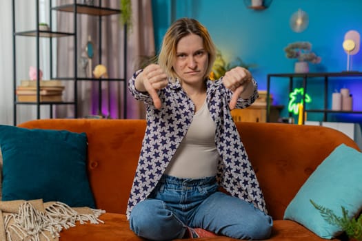 Dislike. Portrait of upset woman showing thumbs down sign gesture, expressing discontent, disapproval dissatisfied bad work at modern home apartment indoors. Displeased girl in living room on couch