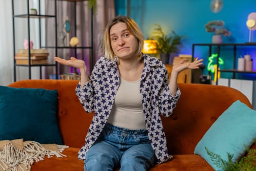 I don't know what to say. Confused Caucasian woman feeling embarrassed about ambiguous question having doubts, no idea, being clueless and uncertain. Blonde girl sitting on sofa in living room at home