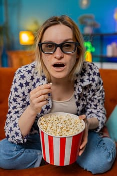 Excited woman sitting on couch eating popcorn watching interesting TV serial, sport game, film, online social media movie content at home. Girl in 3D glasses enjoying domestic entertainment. Vertical
