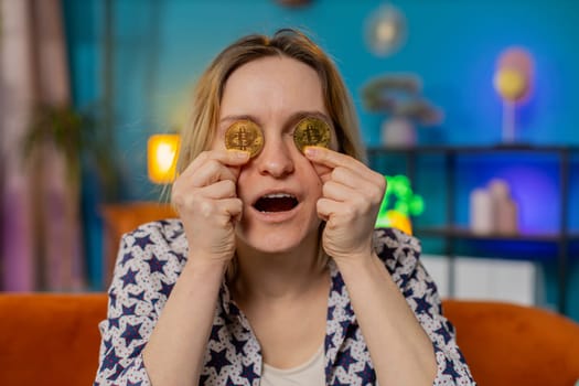 Portrait of happy woman holds two gold BTC coins at home successful developer programmer. Girl stock trader earning bitcoins after online monitoring trading operations. Increasing wealth covering eyes