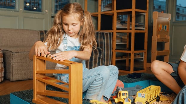 Caucasian girl sits at sand box while placed car model at slope toy. Diverse children playing plastic model while sitting at school in break time at play room. Creative activity concept. Erudition.