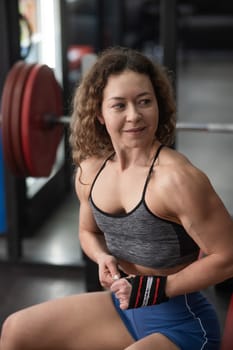 Caucasian forty-year-old woman puts on wrist straps before doing barbell exercises in the gym. Vertical photo