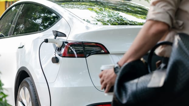 Young man recharge electric car's battery from charging station in outdoor green city park in springtime. Rechargeable EV car for sustainable environmental friendly urban travel lifestyle. Expedient