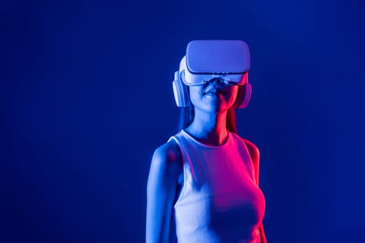 Smart female stand with surrounded by cyberpunk neon light wear VR headset connecting metaverse, futuristic cyberspace community technology. Elegant woman look generated virtual area. Hallucination.