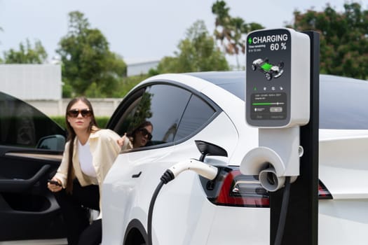 Young woman recharge her EV electric vehicle at green city park parking lot. Urban sustainability lifestyle for environmental friendly EV car with battery charging station. Expedient