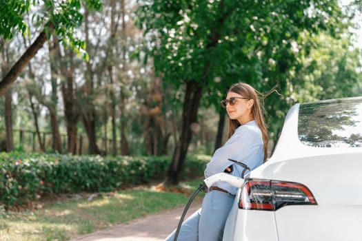 Young woman using smartphone online banking application to pay for electric car battery charging from EV charging station during vacation holiday road trip at national park or summer forest. Exalt