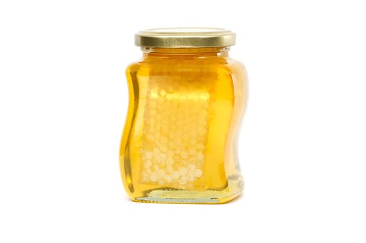 A glass jar with a golden lid contains honey with honeycomb filled almost to the top, against a white background