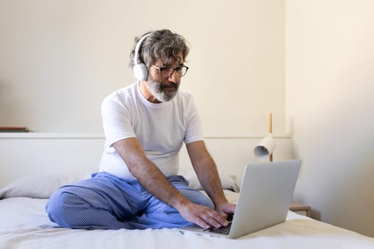 Mature man working, typing on laptop and listening to music with wireless headphones in bed at home. Working from home concept.